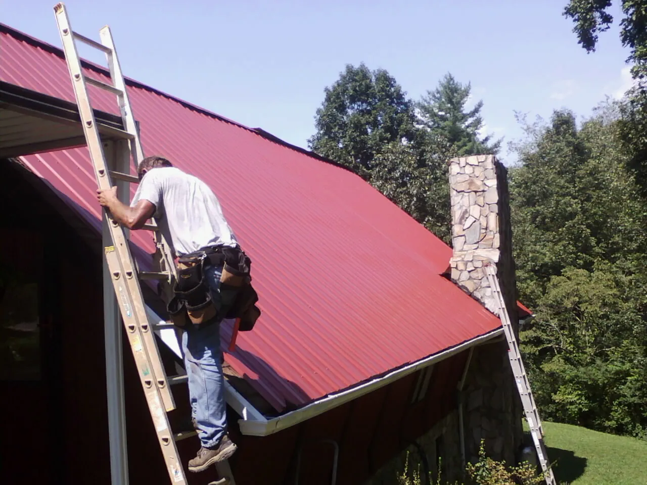 Roofer on a ladder beside a red metal roof and stone chimney — Soffit & Fascia Repair by Roofing Brothers in Haverford